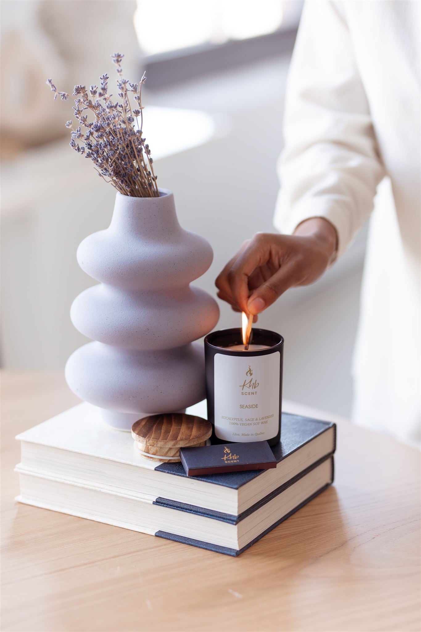 Person lighting a candle using wooden matches for candles next to a decorative vase and books